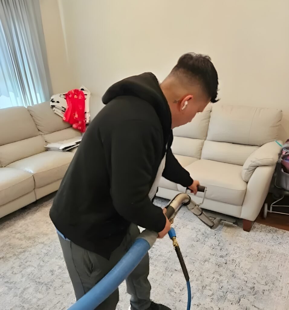 Technician in a black hoodie cleaning a light gray area rug with a wand in front of beige leather sofas