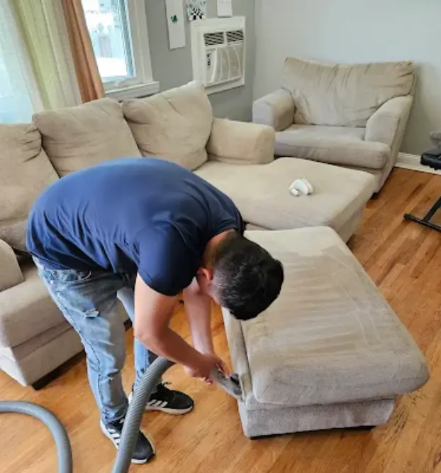 Technician cleaning a beige sofa and ottoman that stand on a wood floor in a bright living room