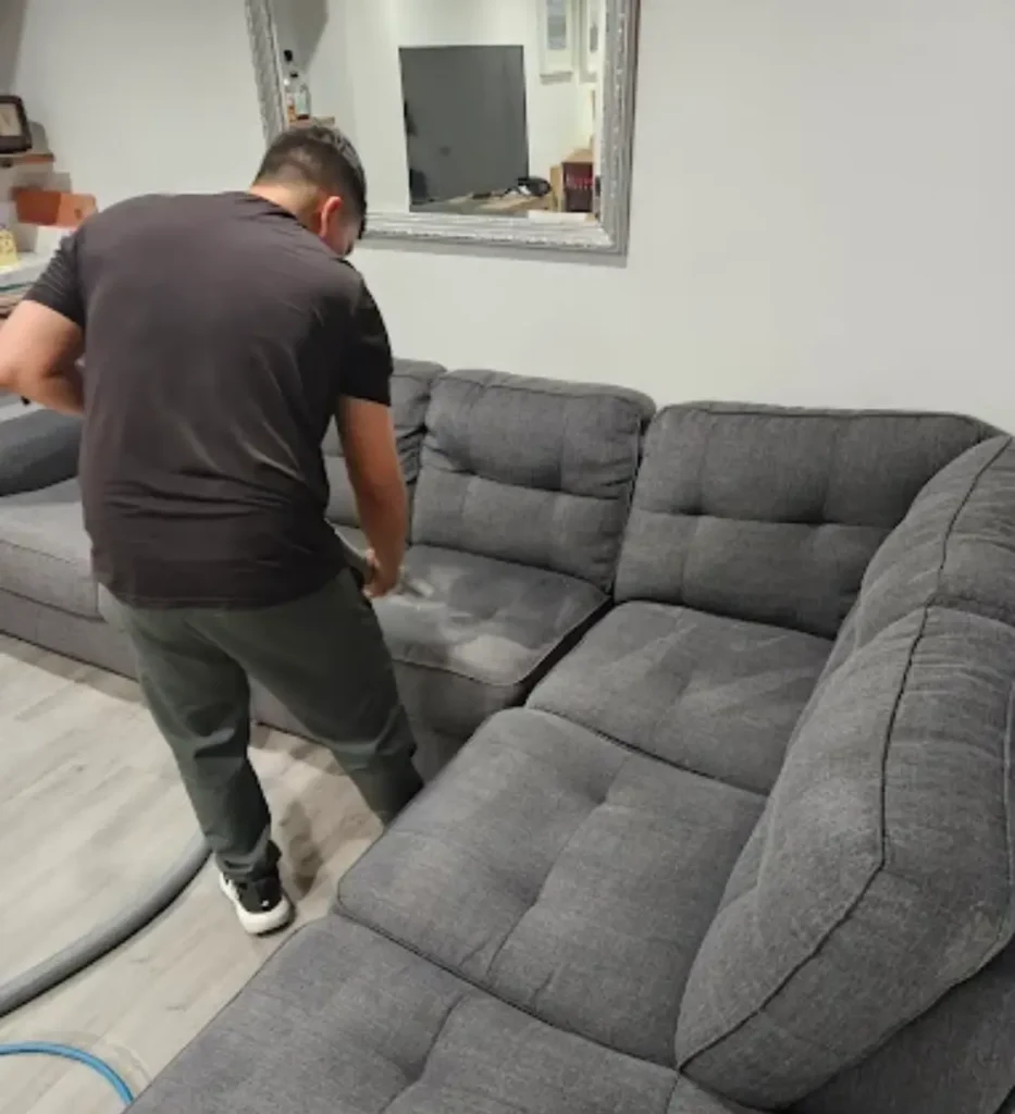 Technician cleaning a large gray fabric sectional sofa in a living room