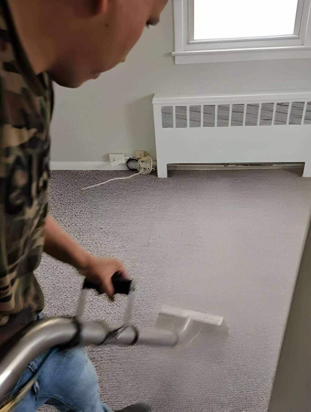 Technician cleaning gray wall-to-wall carpet with a cleaning wand near a white radiator and window